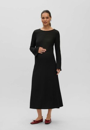 Femme aux cheveux foncés portant une longue robe noire et des ballerines rouges, debout avec une main sur le ventre devant un fond gris uni.