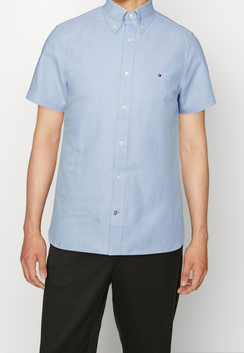 Homme portant une chemise bleu clair à manches courtes avec un petit logo brodé sur la poitrine gauche, associée à un pantalon noir.