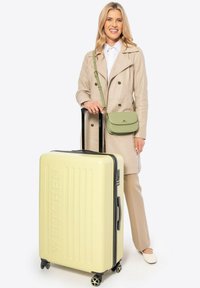 Valigia rigida gialla con scanalature verticali, zip nera e ruote, accanto a una persona in un trench beige che tiene una borsa verde.