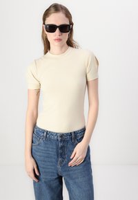 Μπεζ ριμπ τοπ με κοντά μανίκια, φορεμένο με ψηλόμεσο μπλε τζιν. Το μοντέλο έχει αξεσουάρ μαύρα γυαλιά ηλίου και ασημένια σκουλαρίκια κρίκους.