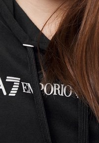 Svart hoodie med vitt logotyp, med snodd och textilstruktur. Närbild på halsringning och hår, som betonar material och färgkontrast.