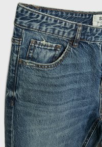 Vaqueros de mezclilla en azul medio con un efecto desgastado. Presentan una cinturilla estándar, un bolsillo frontal y detalles de costura visibles.