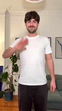 Jaunas vyras su rudais plaukais ir barzda mojuoja dešine ranka svetainėje, kurioje matyti augalai, rėminiai paveikslai ir žalia sofa fone.