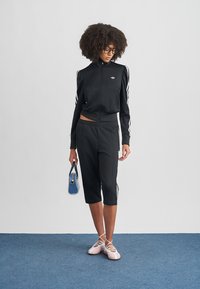 Femme aux cheveux bouclés portant un survêtement noir Adidas, tenant un petit sac à main bleu, et chaussée de ballerines rose avec des brides croisées.
