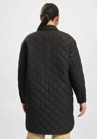 Svart quiltet lang frakk med diamantmønster, myk tekstur, stor krage og forlengede ermer. Har rett hem og sidelommer.