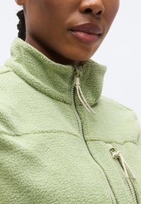 Φλις μπουφάν σε ανοιχτό πράσινο με ψηλό γιακά, μπροστινό φερμουάρ και λεπτομέρεια με κορδόνι. Υφή υφάσματος με ορατές ραφές και ραφές.