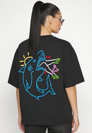 Femme aux longs cheveux noirs et lunettes de soleil porte un t-shirt noir avec un motif coloré représentant un requin mordant les jambes d'une personne et un soleil en arrière-plan.