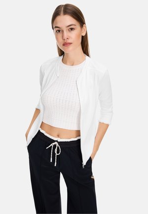 Weiße Zip-Jacke mit gekürzten Ärmeln, getragen über einem weißen strukturierten Top und kombiniert mit schwarzen Jogginghosen mit Kordelzug und Rüschensaum.