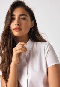 Biela košeľa s kr átkym rukávom a špicatým golierom, hladká textúra, zapínanie na gombíky; model drží ruku pri tvári, čím zdôrazňuje prirodzené črty.