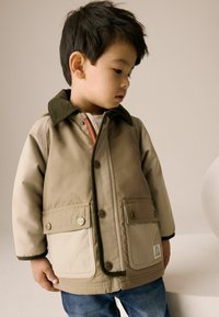 Giacca impermeabile beige e color sabbia con colletto in corduroy, due tasche frontali e bottoni a pressione. Indossata sopra una camicia bianca e jeans blu.