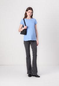 Top scurt ribat de culoare albastru deschis, cu un tiv zimțat, purtat cu blugi negri evazați și o poșetă neagră. Modelul stă pe un fundal neutru.
