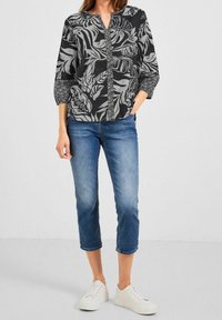 Blusa a fantasia nera e grigia con scollatura a V, maniche a tre quarti e chiusura frontale con bottoni, abbinata a jeans blu cropped e sneaker bianche.