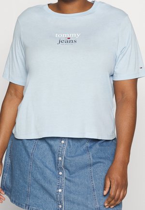 Osoba ubrana w jasnoniebieską krótka koszulkę Tommy Jeans i niebieską spódnicę jeansową z guzikami z przodu, widoczna od szyi do połowy uda.