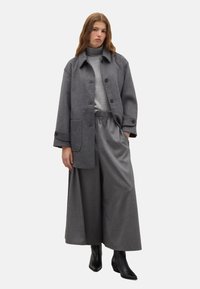 Cappotto grigio in misto lana con grandi bottoni e tasche laterali, abbinato a pantaloni larghi grigi con cintura elastica e stivaletti neri.