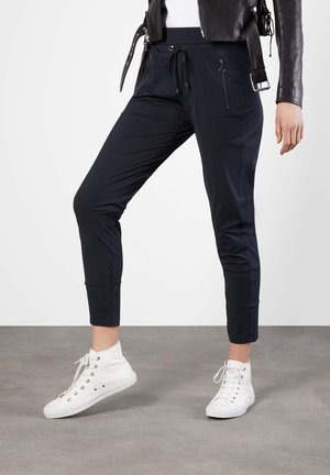 Mujer con pantalones ajustados negros con bolsillos delanteros con cremallera, chaqueta de cuero negra y zapatillas blancas de caña alta, de pie sobre un suelo gris.