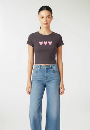 Donna con capelli scuri raccolti in una coda di cavallo, che indossa una maglietta nera corta con tre cuori rosa e jeans a gamba larga, chiari e a vita alta.