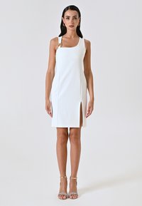 Robe blanche sans manches avec un décolleté carré, fente latérale et un accent doré sur la bretelle gauche ; texture lisse et design ajusté.