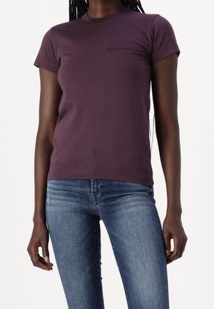 Woman wearing a fitted purple T-shirt and blue jeans, with long braided hair, standing against a plain white background.