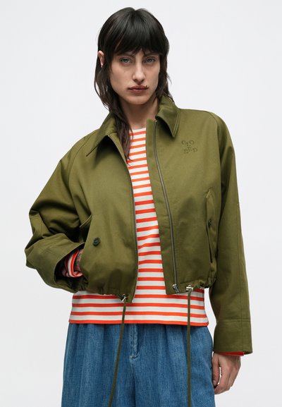 Πράσινο ελιάς cropped μπουφάν με ψηλό γιακά, φερμουάρ μπροστά και λεπτομέρειες με κουμπιά. Φορεμένο πάνω από κόκκινο και λευκό ριγέ πουκάμισο. Casual εφαρμογή.