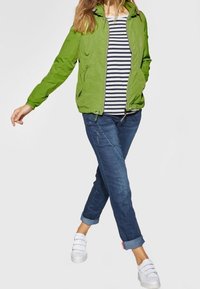 Zielona kurtka z zamkiem i kapturem, noszona na poziomo paskowanym topie. Dżinsy w kolorze niebieskim z podwiniętymi nogawkami oraz białe buty slip-on dopełniają outfit.