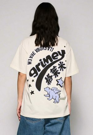 Person mit langen dunklen Haaren trägt ein beige übergroßes T-Shirt mit dem Schriftzug "grimey", chinesischen Schriftzeichen, Sternen und einer Grafik eines geflügelten Bären auf der Rückseite.
