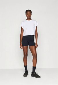 Balts īss krekls ar īsām piedurknēm, kombinēts ar melniem šortiem. Modelis valkā melnas zeķes un apavus, attēlots uz vienkārša fona.