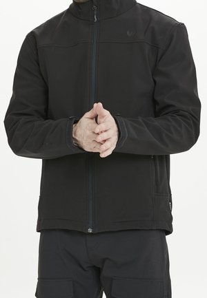 Homme portant une veste noire zippée et un pantalon noir, les mains jointes devant à hauteur de la taille sur un fond uni.