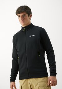 Giacca in pile nera con colletto alto, tasche con zip e cerniere verdi neon a contrasto. Presenta un logo sul lato sinistro del petto.