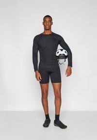 Schwarzes, langärmliges Sportoberteil und Shorts mit enganliegendem Design. Hält einen schwarz-weißen Helm und steht vor einem grauen Hintergrund.