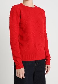 Pull rouge en tricot avec motifs de cœurs en relief, col rond et manches longues. Poignets et ourlet côtelés pour une coupe ajustée.
