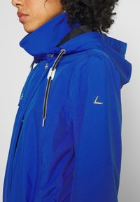 Femme portant une veste imperméable bleue avec un col montant, des cordons noirs et blancs, et un logo métallique sur la manche gauche.