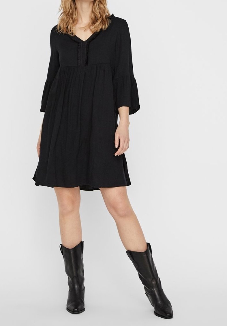 Femme portant une robe noire ample à manches trois-quarts et des bottes en cuir noires mi-mollet, debout devant un fond uni.
