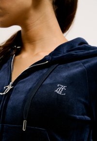 Navyblauwe jumpsuit met capuchon in zachte stof en zilverkleurige hardware. Kenmerkt zich door een strass-logo op de borst en een ritssluiting aan de voorkant.