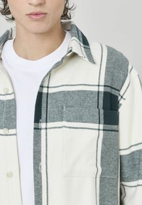 Chemise en plaid en tissu doux, avec un motif à carreaux verts et blancs, devant boutonné et poche poitrine. Design de col classique.