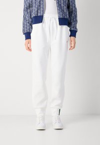 Vita sweatpants med elastisk midjeband och muddar, tillverkade av mjukt material. Kombinerat med en blå mönstrad jacka och vita sneakers.