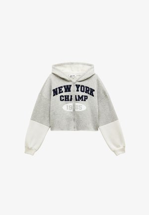 Grijze cropped hoodie met marineblauwe tekst "NEW YORK CHAMP 1985." Heeft witte mouwen en een ritssluiting aan de voorkant. Zachte stof met een capuchon.