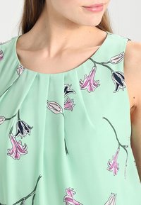 Top senza maniche verde menta con motivi floreali neri e rosa, con dettagli plissettati sul colletto per una maggiore texture.
