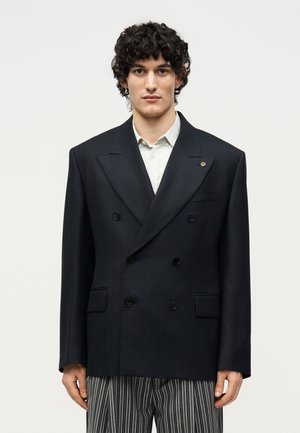 Young man with curly hair wearing an oversized black double-breasted blazer, white shirt, and black-and-white striped pants.