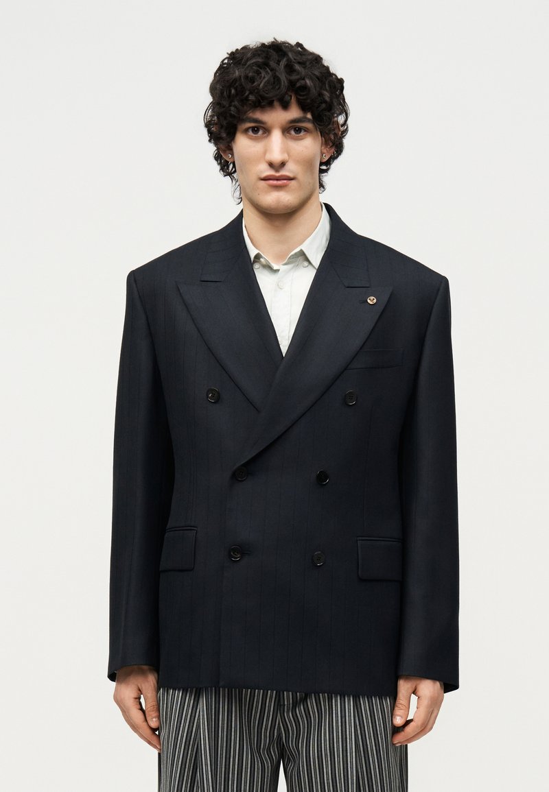 Νέος άνδρας με κατσαρά μαλλιά που φοράει υπερμεγέθη μαύρο διπλό ριγέ σακάκι, λευκό πουκάμισο και μαύρο-λευκό ριγέ παντελόνι.