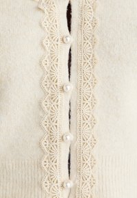 Kremfarget cardigan med intrikate blonder og to perleknapper i midten. Mykt stoff med en teksturert finish.