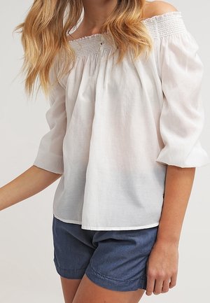 Femme portant un chemisier blanc épaules dénudées avec un col smocké et des manches trois-quarts assorti à un short bleu foncé.