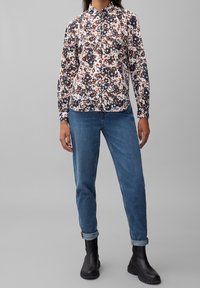 La camicia a maniche lunghe a fantasia floreale presenta un design bianco, blu navy e arancione con colletto a punta. Abbinata a jeans blu con risvolto e stivali neri.