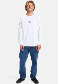 Long-sleeve white shirt with a blue logo, paired with blue denim jeans. Model stands casually, wearing black slip-on shoes.