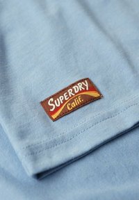 Ljusblå bomulls-tshirt med en brun etikett som står "Superdry Calif." Vita och gula detaljer på etiketten, slät textur, vikta kanter synliga.