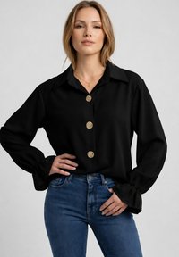 Mujer con blusa negra de manga larga con tres botones dorados, combinada con jeans azules, posando con una mano en la cadera sobre un fondo neutro.