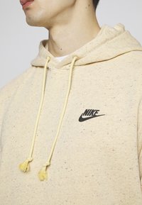 Felpa beige con motivo maculato, caratterizzata da un logo Nike nero. Materiale in misto cotone, con corde di regolazione gialle con nappine.