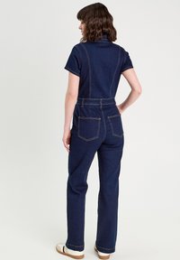 Une combinaison en denim bleu foncé, avec des manches courtes, une taille cintrée et des jambes larges. Coutures contrastantes et deux poches arrière.