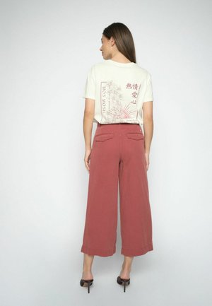 Žena otočená chrbtom, oblečená v krémovom tričku s kvetinovým a textovým motívom, kombinované s vysokými širokými nohavicami v hrdzavej farbe a čiernymi lodičkami.