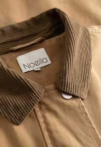 Hnedá košeľa s manžetou z jemného zamatového materiálu a kovovým cvokom. Tkanina je hladká so viditeľným prešívaním a nálepkou s nápisom "Noella."
