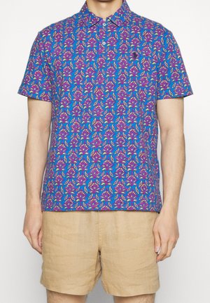 Homme portant un polo à manches courtes bleu avec un motif floral violet et jaune, ainsi qu'un short beige, sur fond blanc.